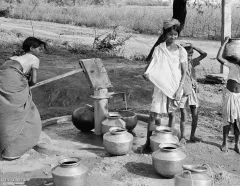 Black-and-white scene of women and children collecting water from a hand pump, carrying metal pots in a rural village; everyday resilience.