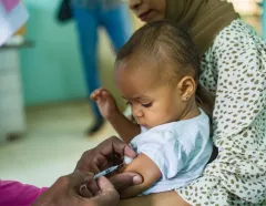 Baby receives a vaccination in the arm while held by a caregiver; a healthcare worker administers the shot in a clinic setting.