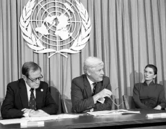 Black-and-white UN meeting: three officials sit at a conference table beneath the United Nations emblem, listening in a formal setting.