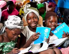 Smiling girls hold bright blue UNICEF books among classmates, while one writes; a lively classroom scene conveys joy and learning.