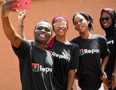 Four smiling adults pose for a selfie outdoors in black Report T-shirts; a sunny red wall and their expressions convey joy and unity.