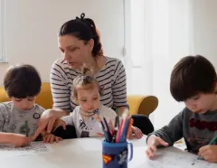 Mother helps two young children draw at a table while a toddler holds colorful pencils; a warm, focused family learning scene.