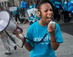 Woman in a UNICEF shirt shouts into a microphone beside a megaphone at a rally, surrounded by blue balloons and supporters; urgent, passionate mood.