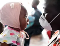 Baby in a pink bonnet and colorful shirt faces a masked caregiver indoors, sharing a curious, tender moment in a warm community setting.