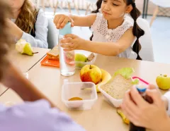 Children share a classroom lunch as a girl pours water into a cup, surrounded by sandwiches, fruit, vegetables, and classmates.